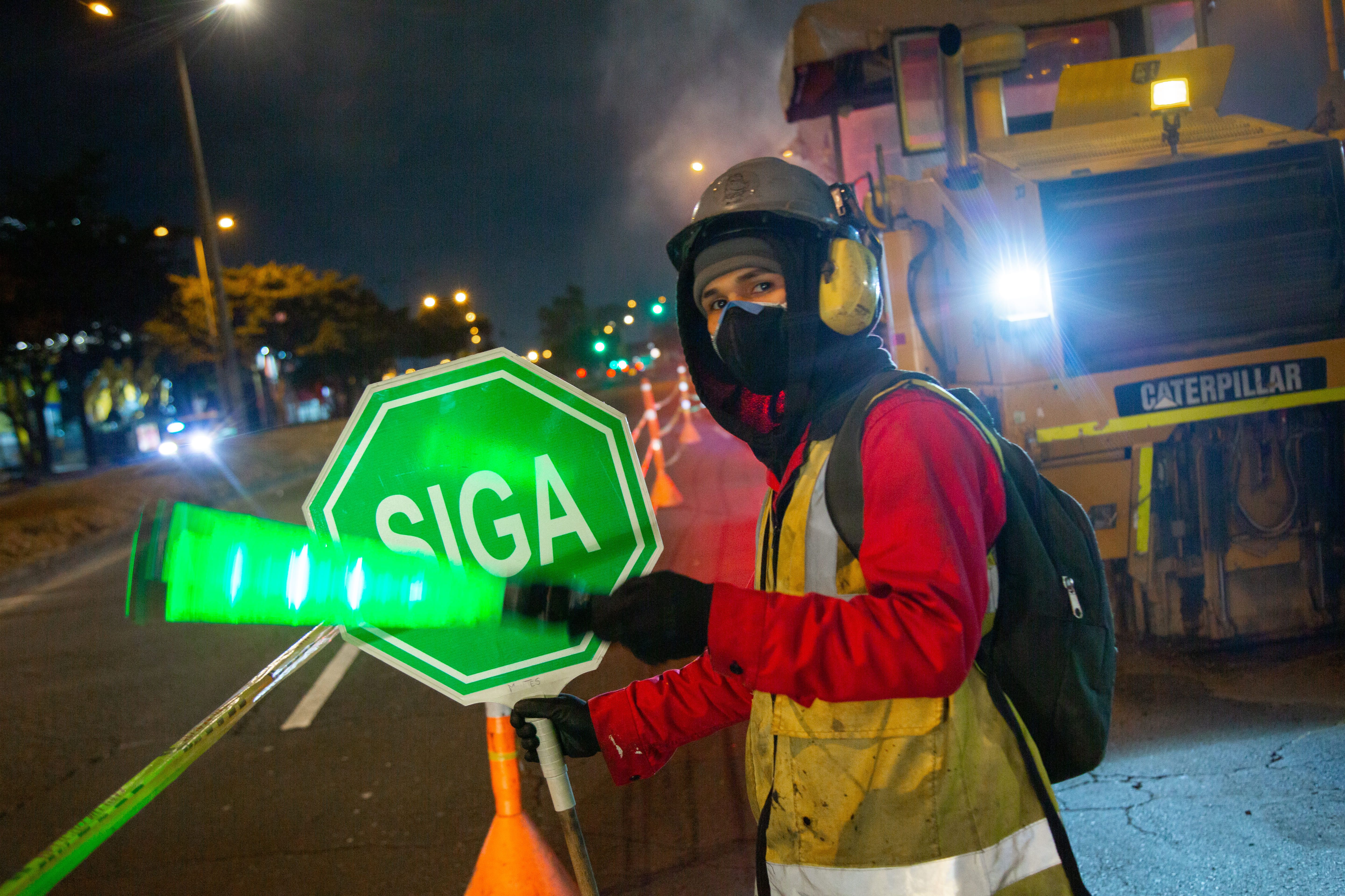 Un joven trabajador de obra sostiene un letrero que dice siga y una paleta fluorecente