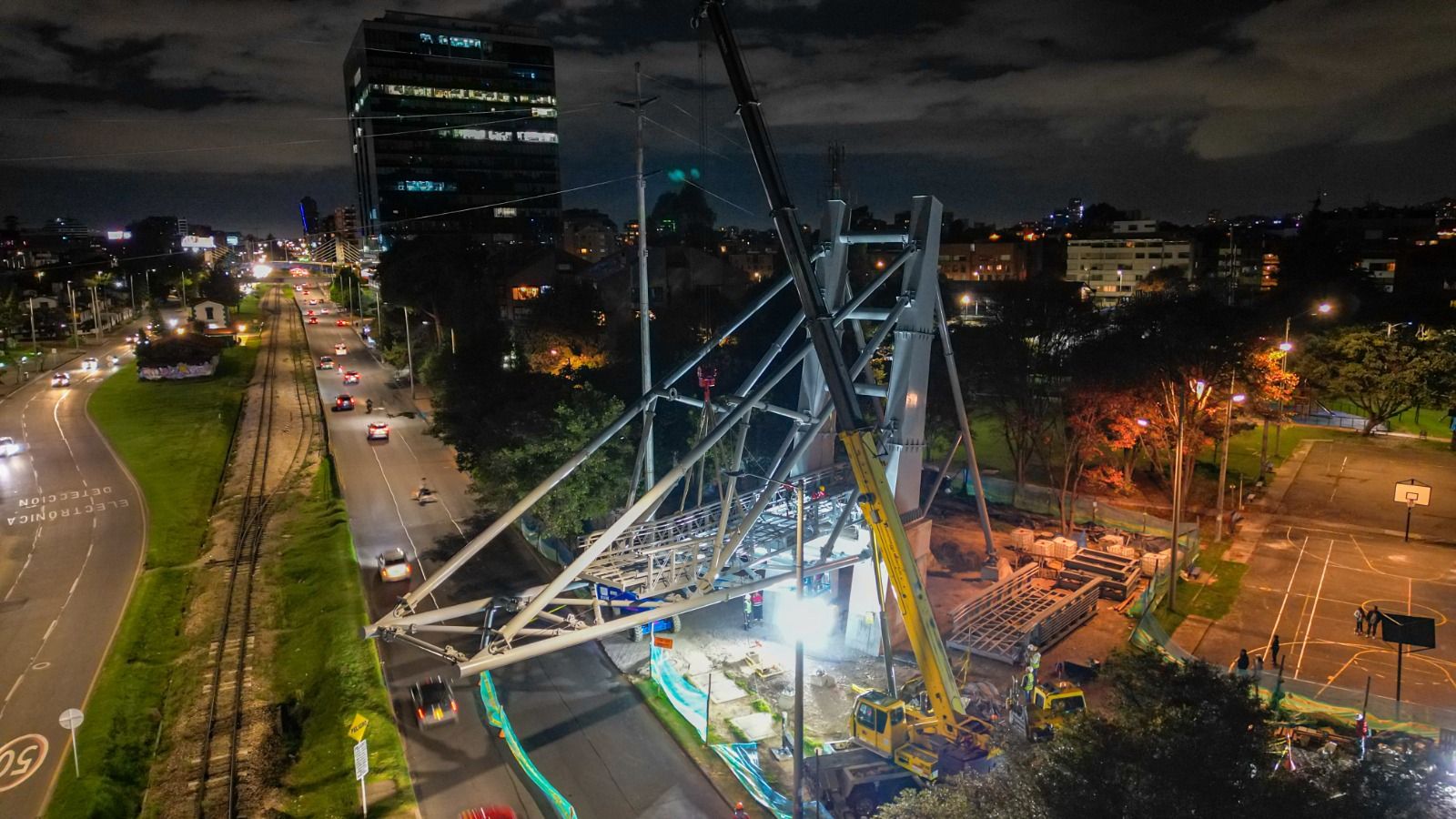 Vista aérea nocturna de una grúa izando e instalando la estructura metálica del puente peatonal sobre la carrera Novena.